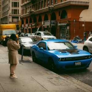 City street view featuring a blue Dodge Challenger parked by the sidewalk amidst urban hustle.