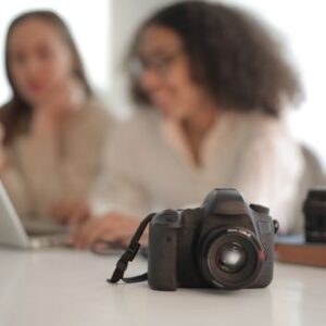 Modern reflex camera with strap placed on desk on background of busy multiethnic women and lenses