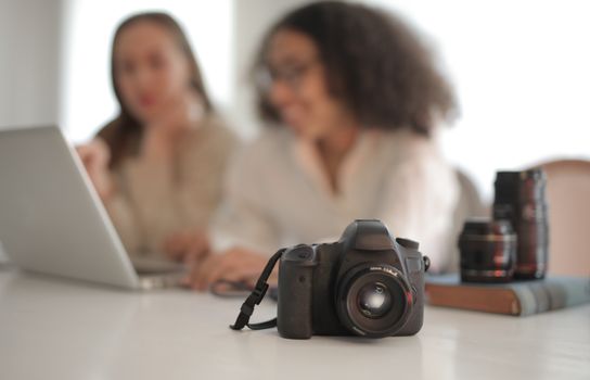 Modern reflex camera with strap placed on desk on background of busy multiethnic women and lenses