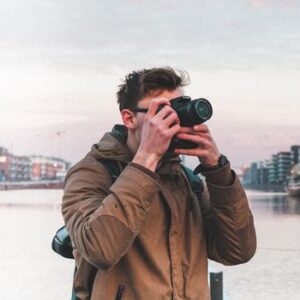 A photographer in a brown jacket takes photos near waterfront with cityscape backdrop.